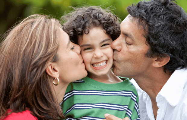 young boy smiling wearing a green and white stripped shirt with his parents both kissing him on the cheek